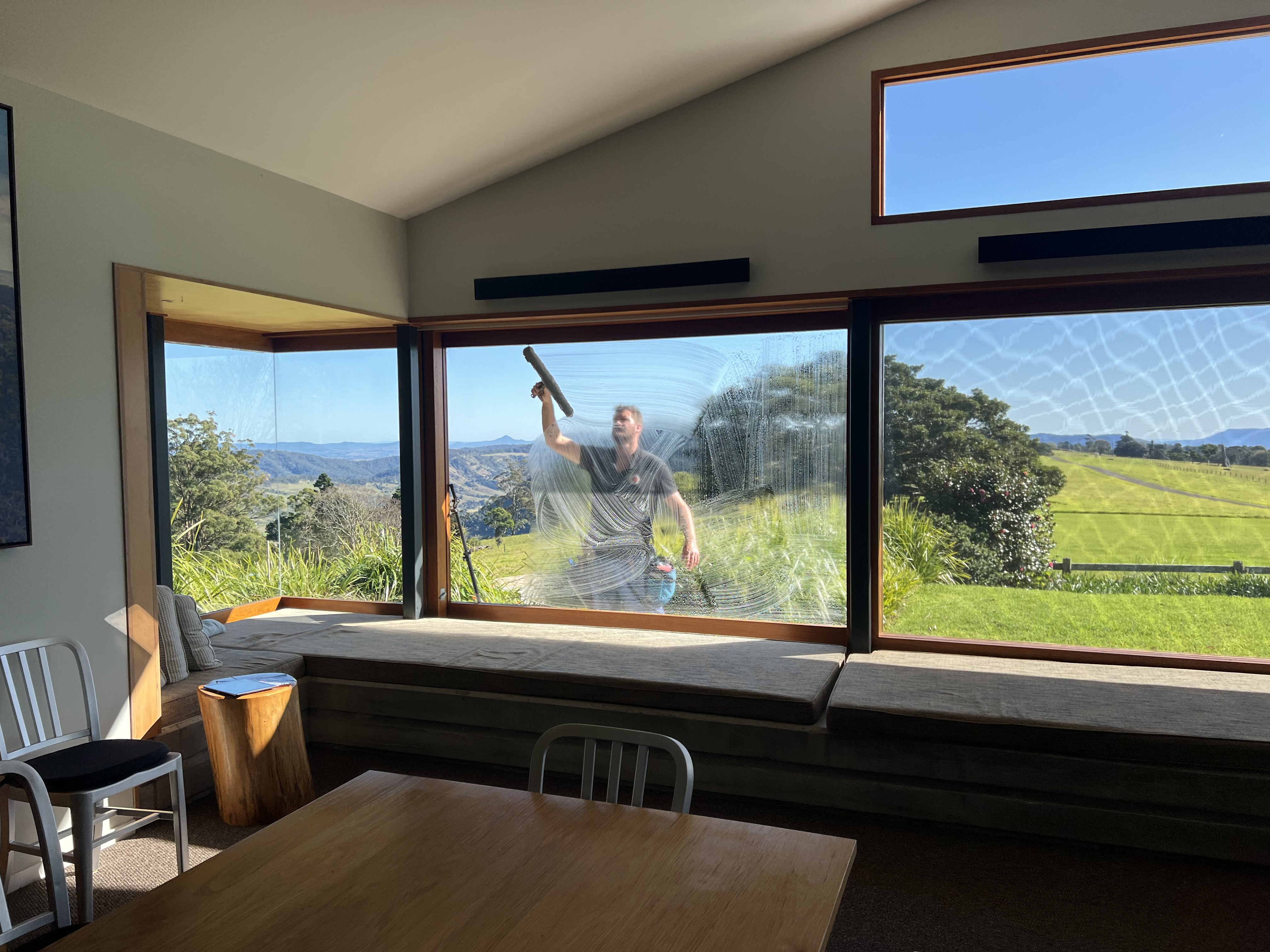 Rhys cleaning a large picture window with a stunning Gold Coast hinterland view of green pasture and mountains beyond the glass