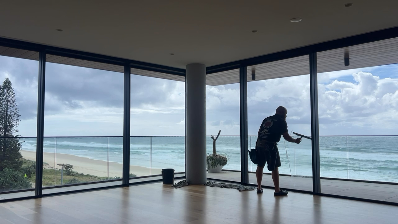 Rhys cleaning floor-to-ceiling windows in a Gold Coast beachfront property — direct ocean views and Zen-branded shirt visible